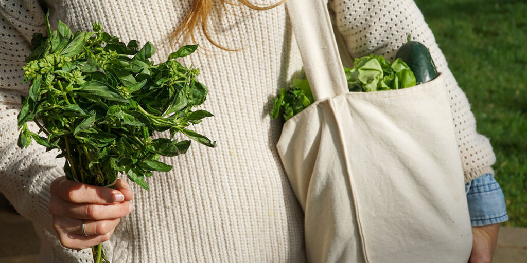 Woman in white sweater returning from the market with a bag filled with leafy greens.
