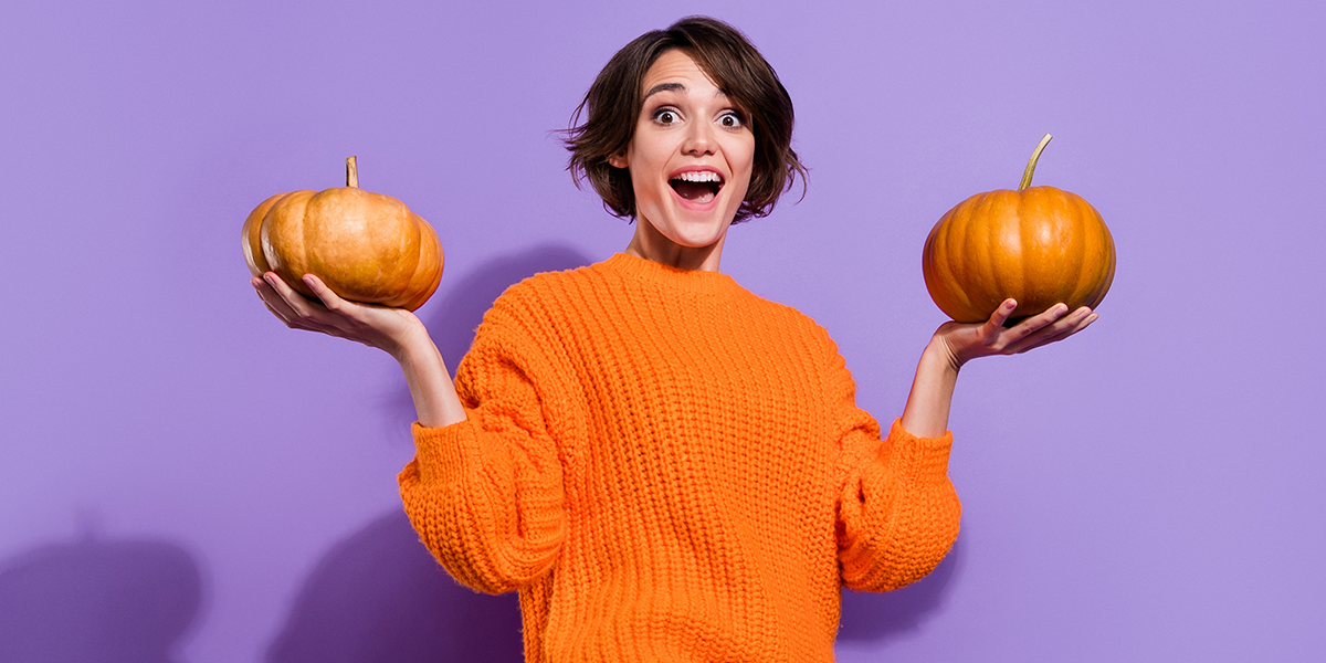An extremely cheerful girl holds two medium-sized pumpkins in her hands against a purple background